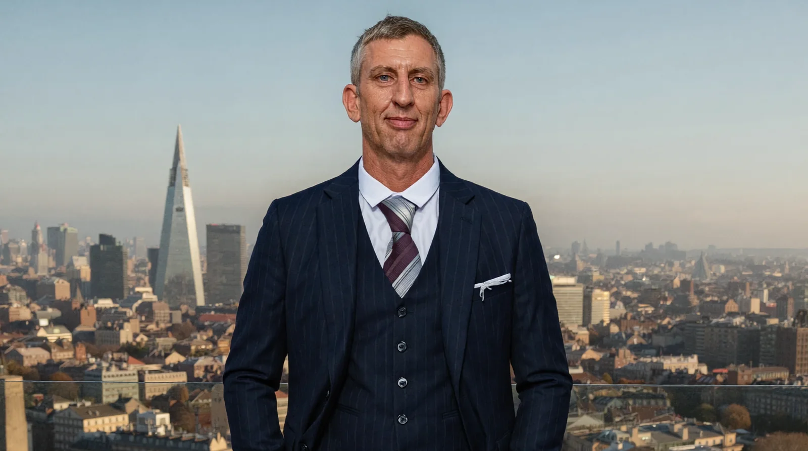 Nick Hall in a three-piece suit on a rooftop terrace with the London skyline and the Shard behind him